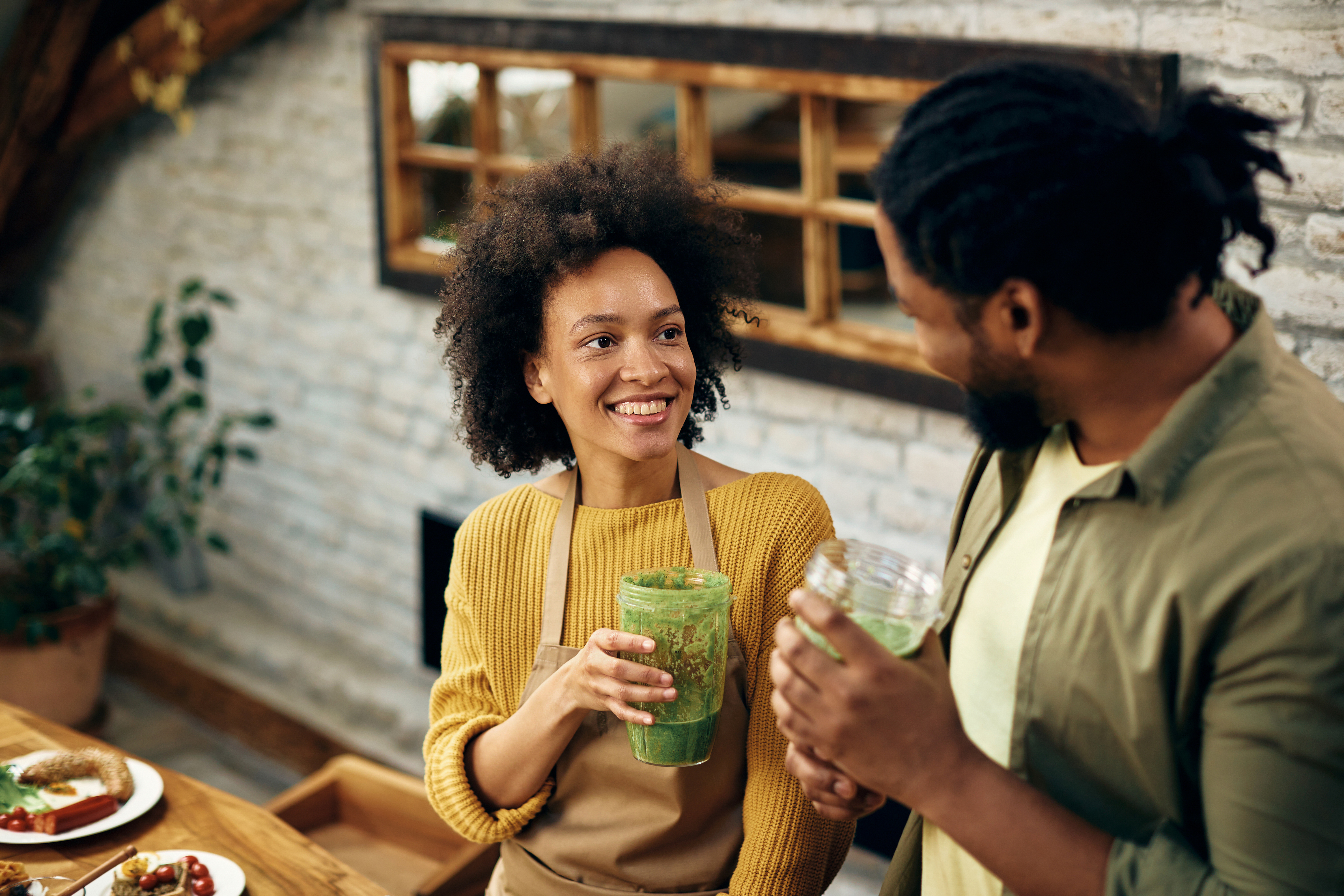 Photo of a Happy couple talking while drinking green smoothie in the kitchen. Focus is on woman.